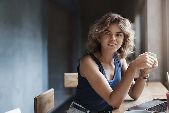 Attracitve modern young woman blond curly hairstyle turn away intrigued smiling delighted hold cup drink coffee working laptop, use net-book studying university task, researching freelance.